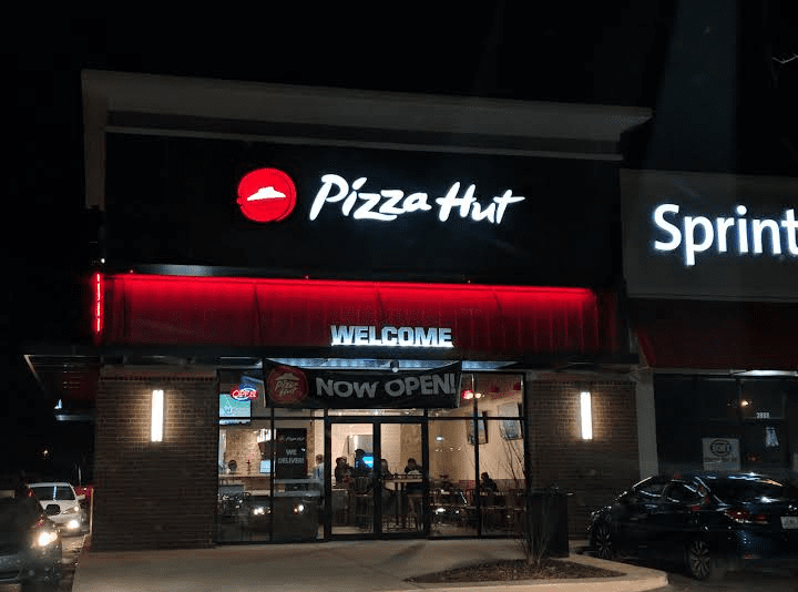 A pizza hut restaurant at night with people sitting outside.
