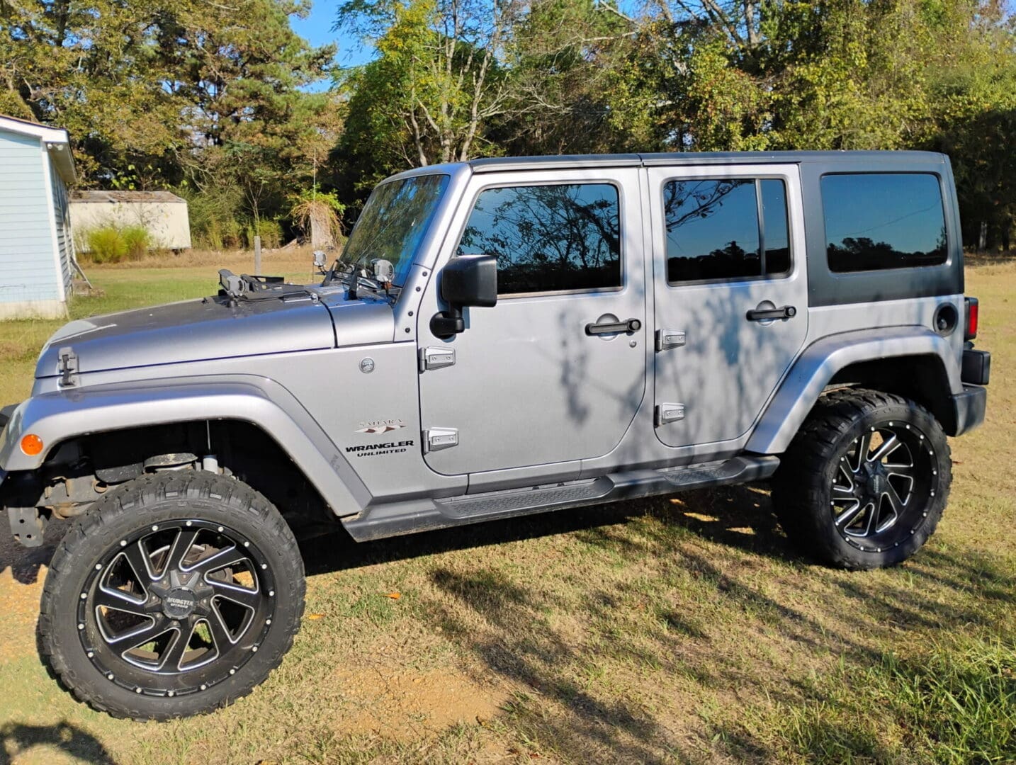 Silver Jeep Wrangler parked on a grassy area with trees in the background.