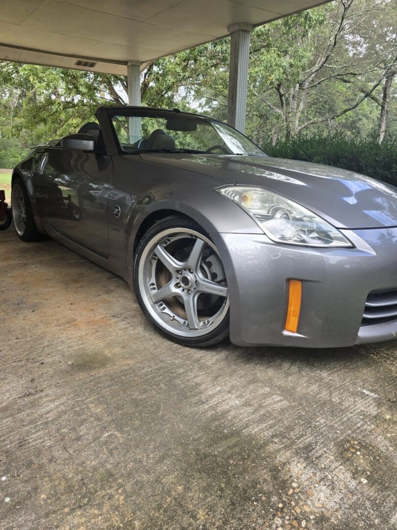 Sleek silver sports car parked under a tree on a driveway.